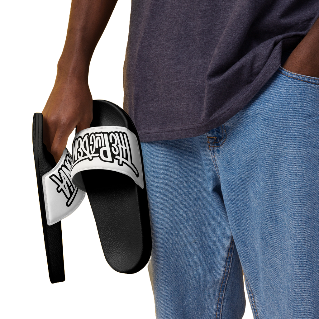 A person wearing a dark gray t-shirt and blue jeans holds a black slide sandal with a white strap featuring the 'The Powerpuff Girls' logo in black.