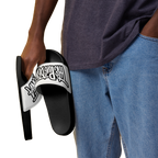 A person wearing a dark gray t-shirt and blue jeans holds a black slide sandal with a white strap featuring the 'The Powerpuff Girls' logo in black.
